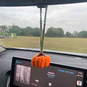 Puede incluir: Un adorno de coche de calabaza naranja hecho a mano, colgado del espejo retrovisor de un coche. La calabaza tiene un tallo marrón y cuerdas de hilo verde. La pantalla del sistema de infoentretenimiento muestra la canción "august" de Taylor Swift.