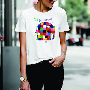 Könnte beinhalten: Weißes T-Shirt mit einem farbenfrohen, geometrischen Elefantenmuster und dem Text "Be yourself". Der Elefant besteht aus Quadraten in Rot, Blau, Grün, Gelb und Schwarz. Das T-Shirt wird mit einer schwarzen Hose getragen.