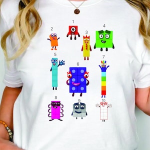 May include: White t-shirt featuring colorful cartoon number characters from 1 to 10. Each number has a unique design and expression, with vibrant colors like red, orange, green, and blue. The shirt is a casual, fun design.