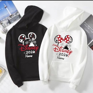 Puede incluir: Dos sudaderas con capucha, una negra y otra blanca, con un diseño de Disney. El diseño incluye orejas de Mickey Mouse, fuegos artificiales, la palabra "Disney" en rojo, el año "2026" y espacio para un nombre. Ambas sudaderas tienen un bolsillo delantero.