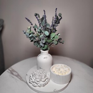 Puede incluir: Un jarrón blanco acanalado con tallos de lavanda y eucalipto secos, acompañado de un joyero en forma de flor y una vela blanca acanalada, todo dispuesto sobre una bandeja circular blanca con un sutil acabado moteado gris.