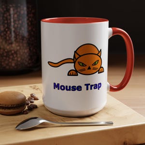 May include: White ceramic mug with a red interior and handle. The mug features an orange cartoon cat illustration with green eyes and the words "Mouse Trap" in blue.