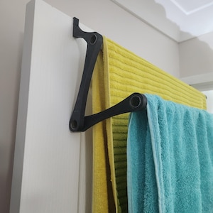 Op de afbeelding: Een zwarte, opvouwbare handdoekhouder aan een witte deur. De houder bevat twee handdoeken: een gele en een turquoise. Het ontwerp maakt het mogelijk om de houder uit te zwaaien voor gebruik en plat op te vouwen wanneer deze niet in gebruik is.