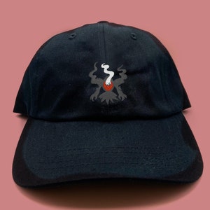 Könnte beinhalten: Eine schwarze Baseballkappe mit einem gestickten Design einer dunklen Kreatur mit einem roten Auge und weißem Rauch, der aus ihrem Kopf kommt.