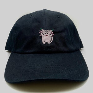 May include: A black baseball cap with an embroidered pink and green cartoon character on the front.