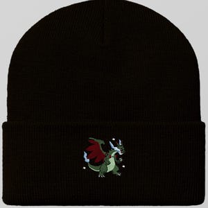 May include: Black beanie hat with an embroidered green dragon design. The dragon has red wings, white horns, and blue accents. The hat is made of a soft, knitted material and has a folded cuff.