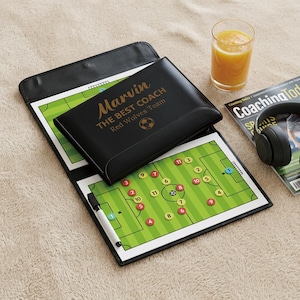 Könnte beinhalten: Ein schwarzes Fußball-Coaching-Klemmbrett mit goldenem Text "Marvin THE BEST COACH Red Wolves Team". Das offene Klemmbrett zeigt ein Fußballfeld-Diagramm mit nummerierten Spielerpositionen. Ein Glas Saft und eine Zeitschrift sind ebenfalls zu sehen.