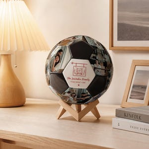 May include: A custom football featuring black panels and white panels printed with personal photographs. One white panel displays a red graphic of a window with the text The Leander Family HOME SWEET HOME. The ball rests on a light wood stand.