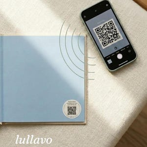 May include: A light blue book with a QR code sticker and a smartphone scanning the code. The book is open, revealing blank pages. The phone is in a green case. The text "lullavo RECORDABLE VOICE STICKERS" is at the bottom.