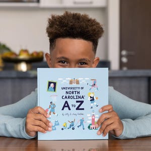 May include: A light blue book titled "UNIVERSITY OF NORTH CAROLINA A to Z" held by a person. The book cover features illustrations of people playing sports and other activities. The book is on a wooden table.