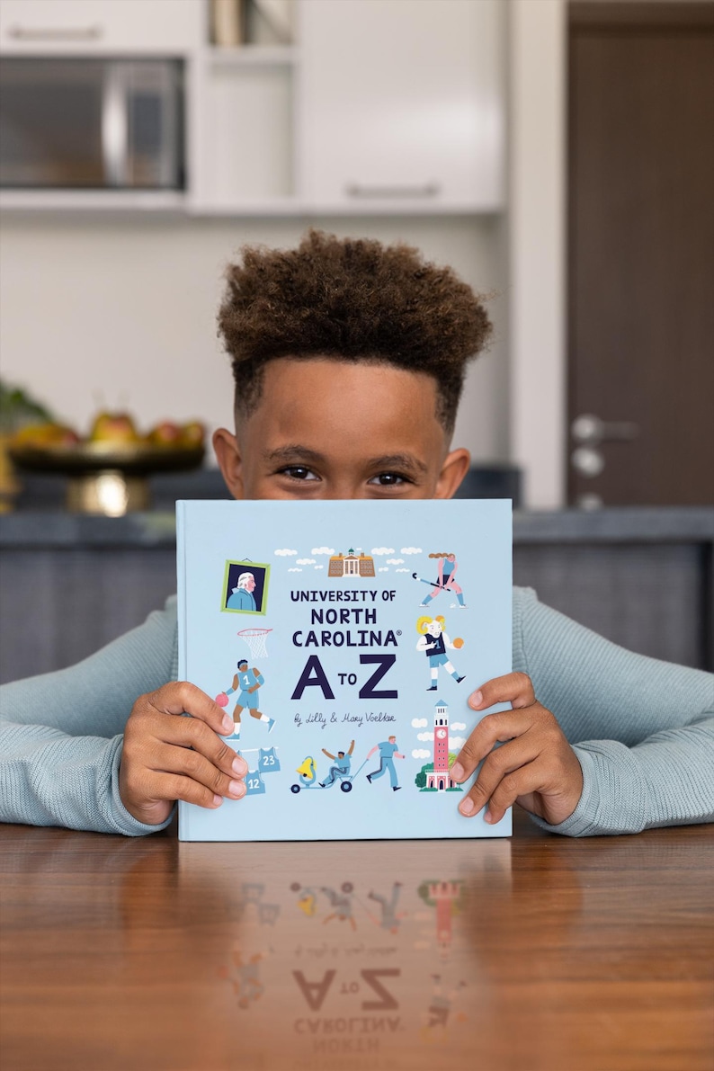 May include: A light blue book titled "UNIVERSITY OF NORTH CAROLINA A to Z" held by a person. The book cover features illustrations of people playing sports and other activities. The book is on a wooden table.