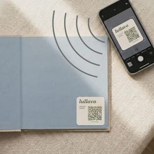 以下が含まれることがあります： 「lullavo」と「SCAN TO LISTEN」という文字がQRコードの上に記された正方形の白いステッカーが貼られた水色の本。近くにはスマートフォンがあり、カメラのインターフェースを通して同じステッカーが曲線とともに表示されている。