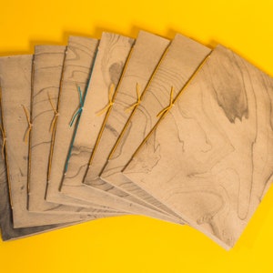 May include: A stack of eight brown paper journals with marbled patterns and tied with yellow string. The journals are arranged in a fan shape on a yellow background.