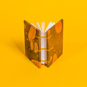 May include: A small, handmade book with a brown cover and orange splotches. The book is open and the pages are white. The book is sitting on a yellow surface.