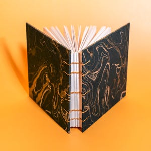 May include: Open handmade journal with a black and gold marbled cover. The pages are white, and the binding is secured with gold thread. The journal is displayed open against an orange background.