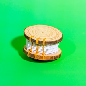 May include: A wooden spool with white thread wrapped around it, secured with orange rubber bands. The spool is sitting on a green surface.