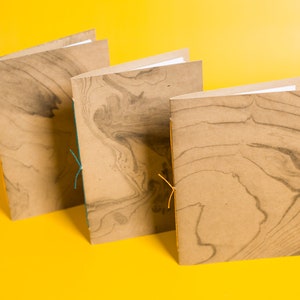 May include: Three brown notebooks with marbled paper covers and orange and teal string closures. The notebooks are arranged in a row on a yellow surface.