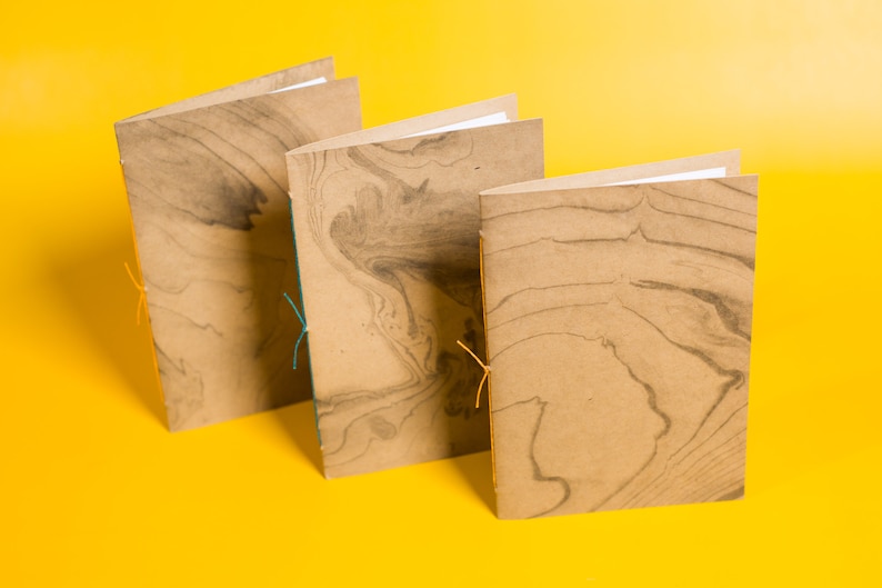 May include: Three brown notebooks with marbled paper covers and orange and teal string closures. The notebooks are arranged in a row on a yellow surface.