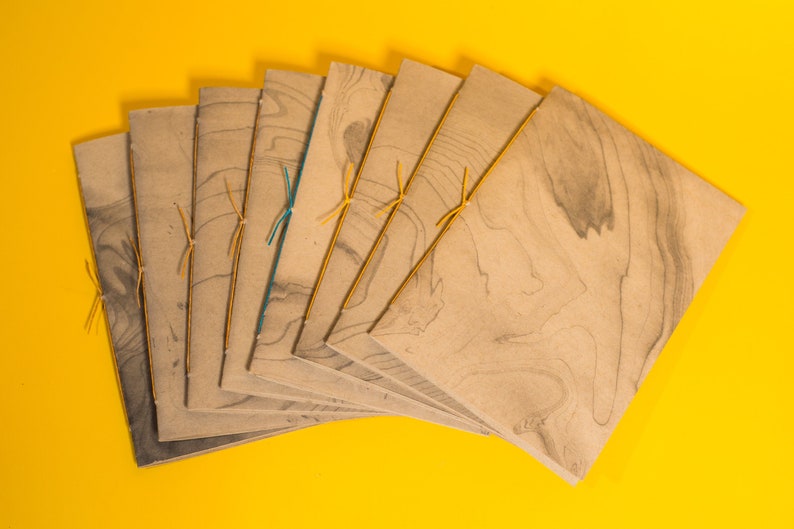 May include: A stack of eight brown paper journals with marbled patterns and tied with yellow string. The journals are arranged in a fan shape on a yellow background.