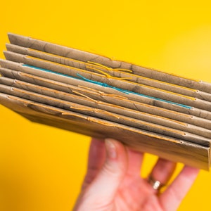 May include: A stack of brown paper notebooks bound with yellow and blue thread. The notebooks are held in a hand.