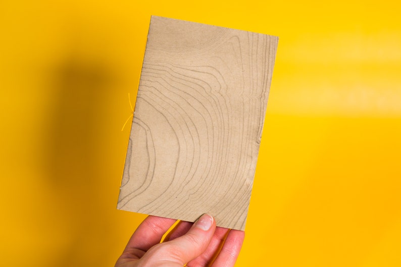 May include: A brown paper notebook with a wood grain pattern. The notebook is held in a hand against a yellow background.