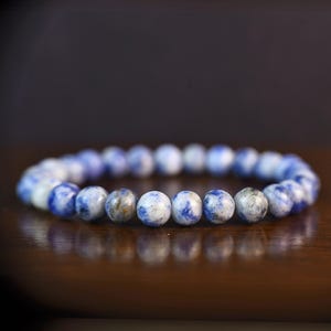 May include: A beaded bracelet featuring round, blue and white speckled beads. The bracelet is resting on a dark wooden surface, with a blurred background. The beads have a marbled appearance.