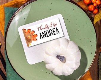 May include: Thanksgiving place setting with a green plate, gold silverware, and a white pumpkin. A place card with a turkey illustration and the name "Andrea" is on the plate. The table is decorated with fall foliage and pumpkins.