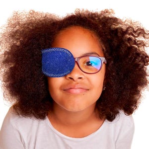 May include: A blue glittery fabric eye patch with white stitched edges, designed to slide over the lens of a pair of glasses. The patch features a textured, sparkling surface and a contoured shape that covers one eye while attached to the frame.