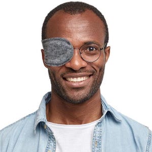 May include: A grey felt eye patch with dark stitching around the edges, designed to fit over one lens of a pair of thin-rimmed metal glasses. The patch is oval-shaped and covers the right eye, featuring a textured fabric surface and visible seams.