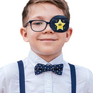 May include: A dark blue denim eye patch featuring a yellow embroidered star with eyes, attached to black-rimmed glasses. The ensemble includes a navy blue bow tie with a small gold star pattern and matching navy blue suspenders over a white shirt.