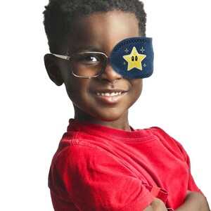 May include: A navy blue fabric eye patch featuring an embroidered yellow star with eyes and small gold sparkles. The patch is designed to slide over the lens of a pair of glasses, covering one eye with a stitched border detail.