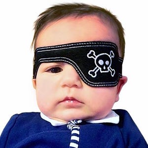 May include: A black fabric eye patch featuring a white embroidered skull and crossbones design. The patch is secured with a matching black strap that wraps around the head, with visible white stitching along the edges of the patch and the strap.