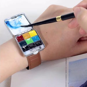 Puede incluir: Un juego de pintura de acuarela en miniatura en una caja de metal plateado, usado en la muñeca con una correa de cuero marrón. El juego incluye una variedad de pinturas coloridas y un área de mezcla pequeña. Un pincel se sostiene sobre el juego de pintura.