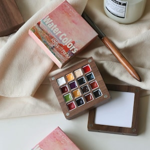 May include: A watercolor paint set with a wooden palette containing 16 colorful paint squares. The set includes a box with the words "Water Colors 1864-1930" and a wooden-handled brush. The palette is open, revealing the vibrant colors.