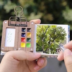 May include: A miniature watercolor painting kit with a wooden palette, a metal clip, and a small sketchbook. The palette holds various colors of paint. The sketchbook displays a watercolor painting of a tree. The kit is held in a person's hand.
