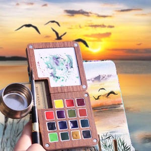 Könnte beinhalten: Ein hölzernes Aquarellfarbenset mit einer Vielzahl von bunten Farben. Das Set enthält eine kleine Mischfläche und einen Pinsel. Der Hintergrund zeigt einen Sonnenuntergang über dem Wasser mit fliegenden Vögeln.