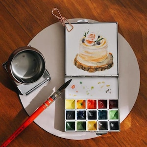 Puede incluir: Un juego de pintura de acuarela con una pintura de pastel en la tapa. El juego incluye un pincel con mango rojo, un recipiente de agua de metal y una paleta de varios colores. Un clip de papel color cobre sujeta la tapa abierta.