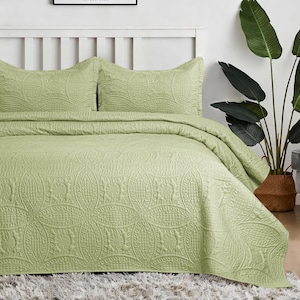 May include: A sage green bedspread and two matching pillow shams featuring a stitched geometric pattern of interlocking circles and textured grid squares. The bedding set is draped over a bed with a white headboard and a shaggy rug.