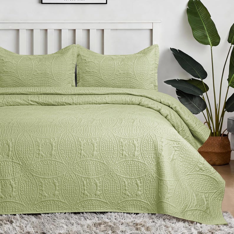 May include: A sage green bedspread and two matching pillow shams featuring a stitched geometric pattern of interlocking circles and textured grid squares. The bedding set is draped over a bed with a white headboard and a shaggy rug.