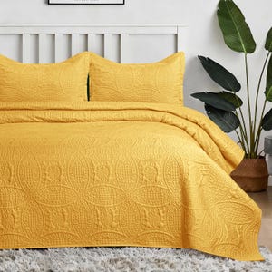 May include: A bright yellow bedspread and two matching pillow shams featuring an embossed geometric pattern of interlocking circles and grid textures. The bedding set is draped over a bed frame with a white headboard and a textured surface.