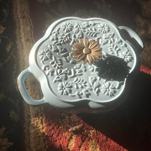 May include: A light blue ceramic serving dish with a floral design on the lid. The lid is adorned with a gold-colored daisy-shaped handle. The dish has two handles and is sitting on a patterned rug.