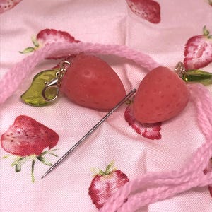 May include: Two red strawberry-shaped charms with silver-colored clasps and a needle rest on a pink fabric with strawberry patterns. A pink cord is looped around the charms. The charms have a textured surface.