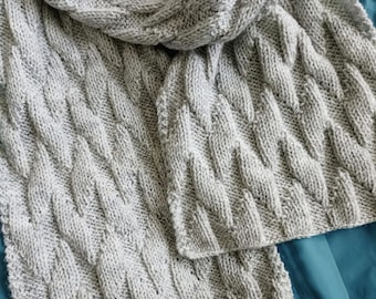 May include: A gray knitted scarf with a cable knit pattern. The scarf is folded and draped over a blue jacket.