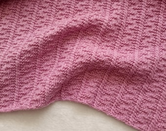 May include: Close-up of a soft, textured, pink knitted blanket. The blanket has a repeating pattern of small, raised rectangles. The fabric is draped over a white, fluffy surface, creating shadows and highlighting the texture of the knit.
