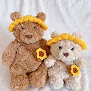 May include: Two plush teddy bears, one brown and one cream-colored, are displayed. Each bear wears a yellow crocheted hat and a brown crossbody bag adorned with a yellow and brown sunflower. The bears are positioned on a white, textured surface.
