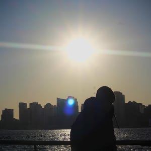 以下が含まれることがあります： 夕暮れの都市のシルエットを背景に、桟橋に立つ人のシルエット。太陽が空に輝き、画像にレンズフレアが発生しています。水は穏やかで反射しています。