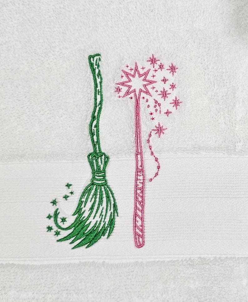 May include: A white towel with green and pink embroidered images of a broom and a wand with stars.