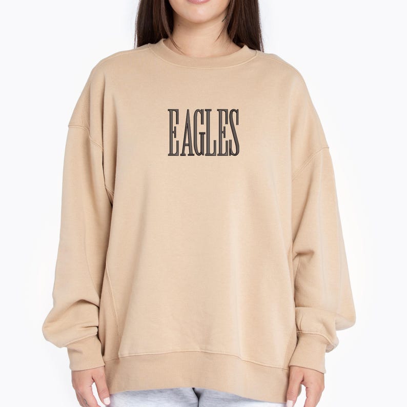 May include: A tan-colored sweatshirt with the word "EAGLES" printed in a black, bold font across the chest. The sweatshirt has long sleeves and a crew neck. The overall style is casual and comfortable.