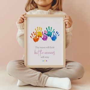 Puede incluir: Un cuadro enmarcado con huellas de manos coloridas en tonos naranja, amarillo, morado, verde y azul. El texto dice: «El desorden se desvanecerá, pero los recuerdos permanecerán». El marco es de color madera clara.