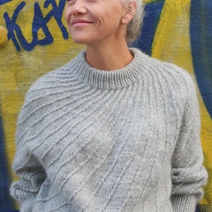 Puede incluir: Un suéter de punto gris claro con un patrón diagonal texturizado. El suéter tiene cuello redondo y mangas largas. El fondo presenta un diseño de graffiti azul y amarillo. La persona de la imagen tiene el pelo gris y está mirando hacia arriba.